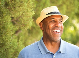 older man in blue polo and fedora smiling amongst trees 